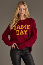 A woman with long blonde hair wears the Panache Apparel Collection's Maroon & Gold Game Day Sweater and black leather pants, standing against a plain gray background.