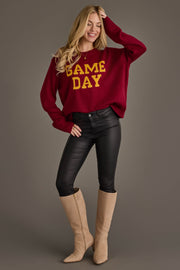 A woman stands and smiles, wearing the Panache Apparel Collection Maroon & Gold Game Day Sweater, black pants, and beige knee-high boots against a plain gray background.