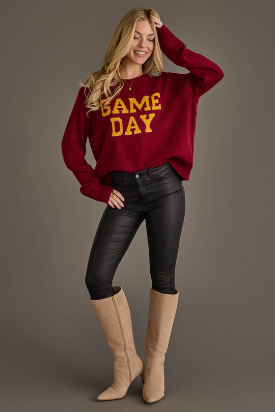 A woman stands and smiles, wearing the Panache Apparel Collection Maroon & Gold Game Day Sweater, black pants, and beige knee-high boots against a plain gray background.