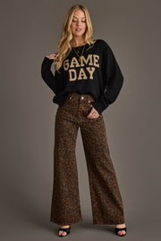 A woman stands against a gray background wearing the Panache Apparel Collection's Black & Tan Game Day Lightweight Sweater, paired with high-waisted brown leopard print wide-leg pants and black open-toed heels.