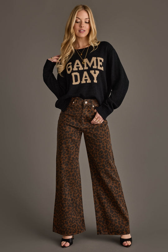 A woman stands against a gray background wearing the Panache Apparel Collection's Black & Tan Game Day Lightweight Sweater, paired with high-waisted brown leopard print wide-leg pants and black open-toed heels.