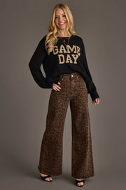 Smiling woman poses with one hand on her hip against a gray background, wearing the Panache Apparel Collection’s Black & Tan Game Day Lightweight Sweater styled with wide-leg leopard print pants and black open-toe shoes.