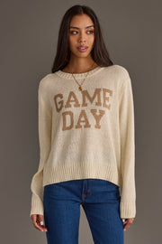 A woman wearing the Ivory Game Day Lightweight Sweater from Panache Apparel Collection, featuring GAME DAY in tan letters, pairs it with blue jeans and gold jewelry as she stands against a plain background, looking slightly to the side.