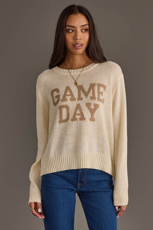 A woman wearing the Ivory Game Day Lightweight Sweater from Panache Apparel Collection, featuring GAME DAY in tan letters, pairs it with blue jeans and gold jewelry as she stands against a plain background, looking slightly to the side.