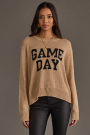 A woman stands against a gray background wearing the Panache Apparel Collection’s Tan Game Day Lightweight Sweater, featuring “GAME DAY” in bold black letters, paired with black pants and gold jewelry.