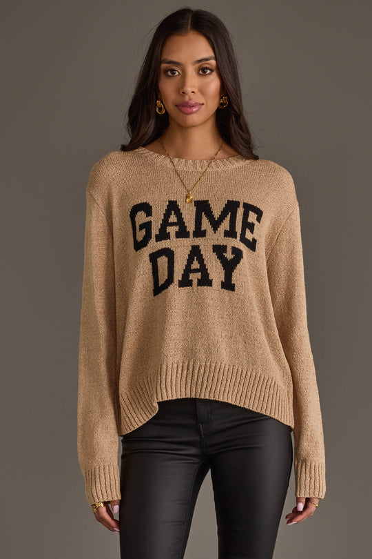 A woman stands against a gray background wearing the Panache Apparel Collection’s Tan Game Day Lightweight Sweater, featuring “GAME DAY” in bold black letters, paired with black pants and gold jewelry.