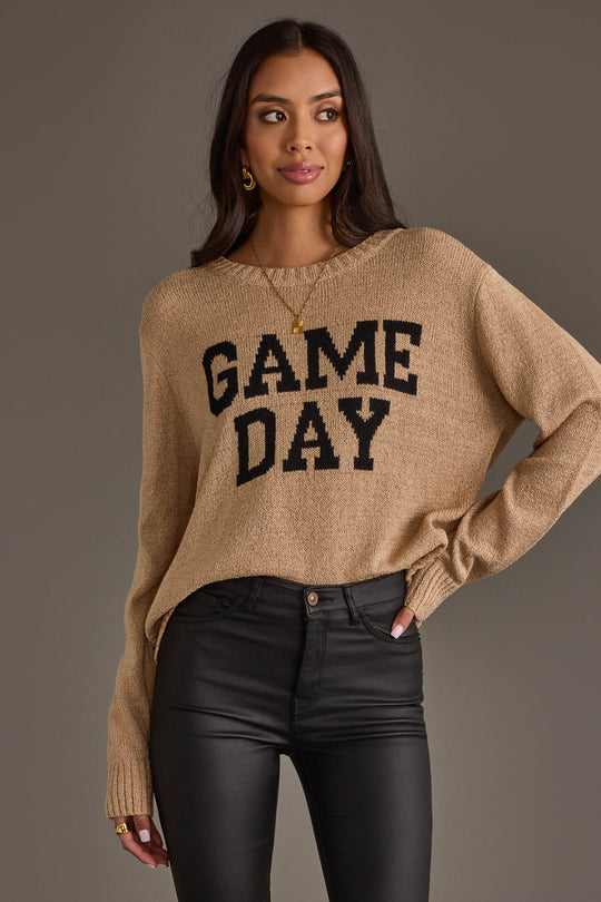 A woman models the Tan Game Day Lightweight Sweater from Panache Apparel Collection, featuring bold black "GAME DAY" lettering, paired with black pants. She stands with one hand on her hip and looks to the side against a plain background.