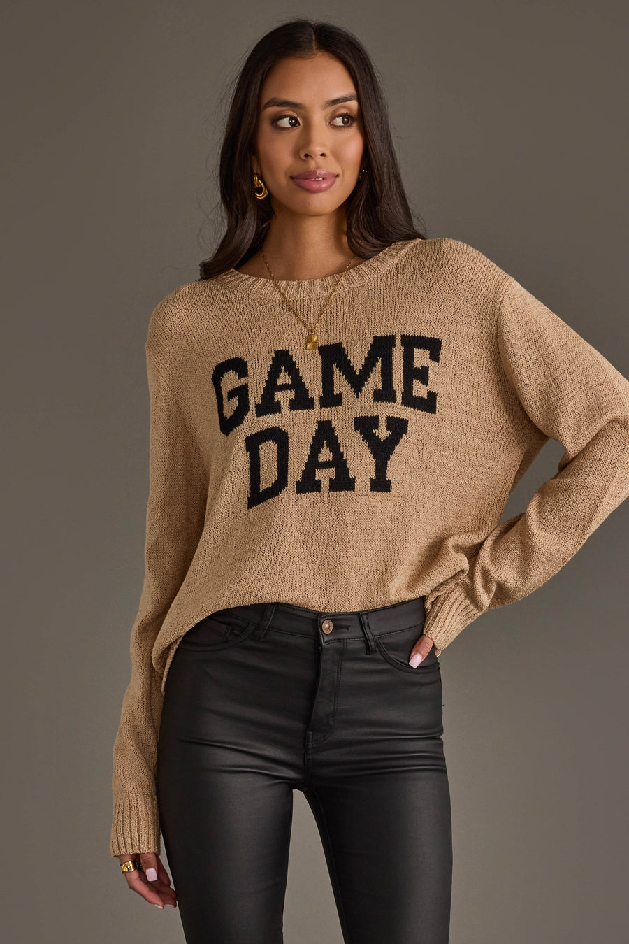 A woman models the Tan Game Day Lightweight Sweater from Panache Apparel Collection, featuring bold black "GAME DAY" lettering, paired with black pants. She stands with one hand on her hip and looks to the side against a plain background.