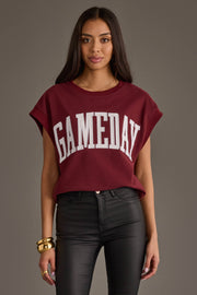 A person wearing a maroon and white "Game Day" short sleeve sweatshirt from the Panache Apparel Collection and black pants stands against a plain background, looking toward the camera. They have long, dark hair and gold jewelry.