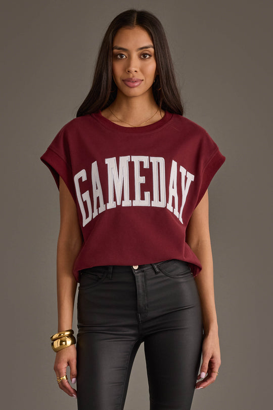 A person wearing a maroon and white "Game Day" short sleeve sweatshirt from the Panache Apparel Collection and black pants stands against a plain background, looking toward the camera. They have long, dark hair and gold jewelry.