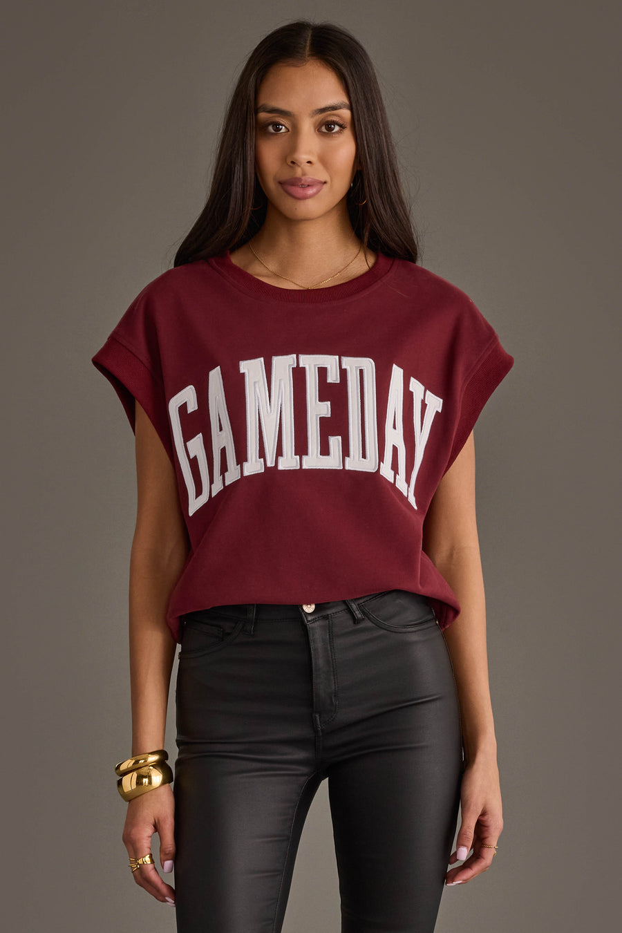 A person wearing a maroon and white "Game Day" short sleeve sweatshirt from the Panache Apparel Collection and black pants stands against a plain background, looking toward the camera. They have long, dark hair and gold jewelry.