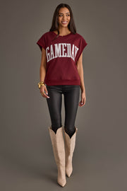 A woman stands smiling against a plain gray background, wearing the Maroon & White Game Day Short Sleeve Sweatshirt by Panache Apparel Collection, paired with black leather pants and cream cowboy boots.