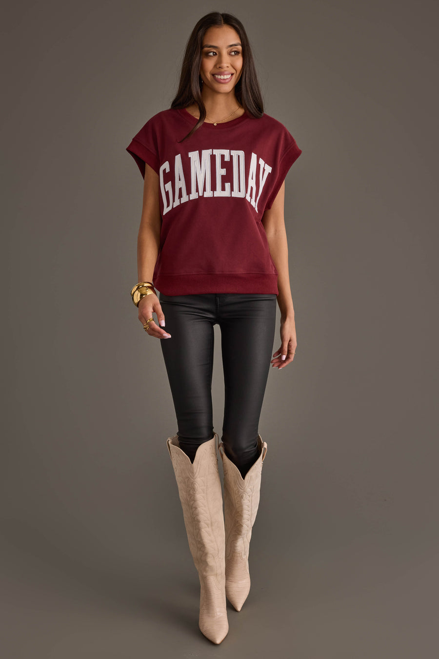 A woman stands smiling against a plain gray background, wearing the Maroon & White Game Day Short Sleeve Sweatshirt by Panache Apparel Collection, paired with black leather pants and cream cowboy boots.