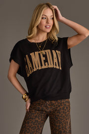 A woman wearing a Panache Apparel Collection Black & Light Brown Game Day Short Sleeve Sweatshirt, brown leopard print pants, gold bracelets, and layered necklaces poses with one hand touching her hair and the other on her hip against a gray background.