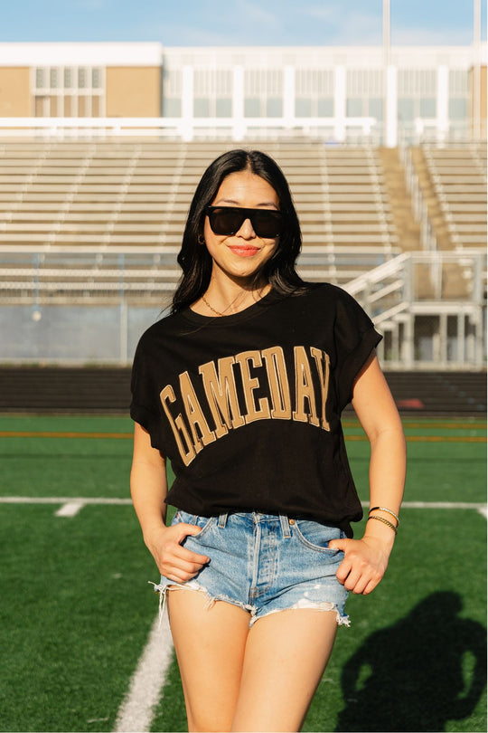 A woman stands on a sports field with bleachers in the background, smiling and wearing sunglasses, denim shorts, and a Black & Light Brown Game Day Short Sleeve Sweatshirt from Panache Apparel Collection.