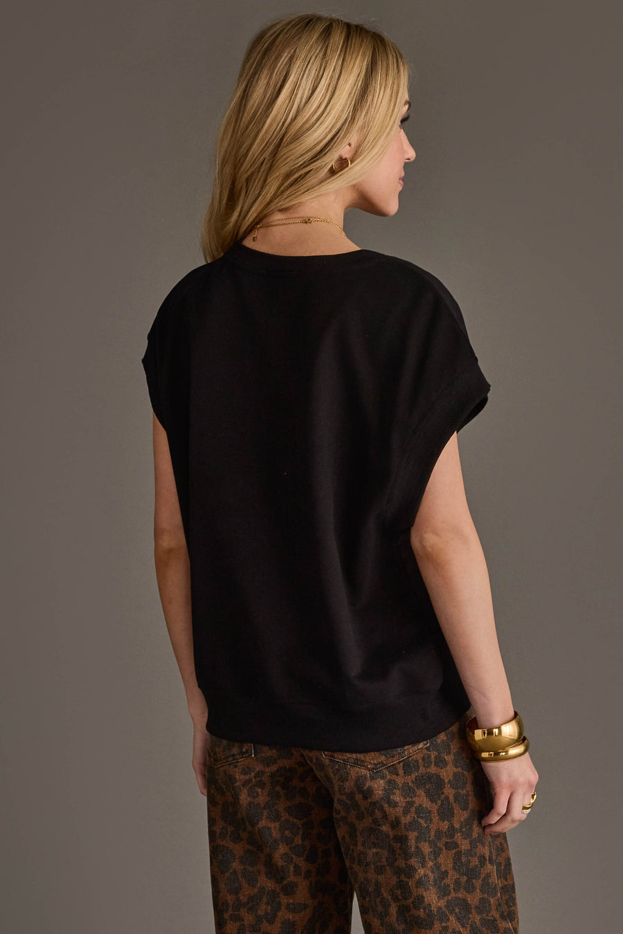 A woman with long blonde hair stands facing away against a plain gray background, wearing the Panache Apparel Collection’s Black & Light Brown Game Day Short Sleeve Sweatshirt and several gold bracelets.