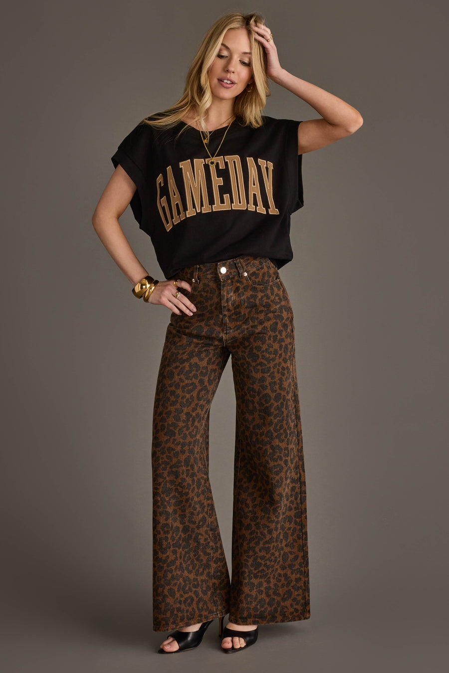 A woman stands against a plain background, smiling slightly with one hand on her hip, wearing a Black & Light Brown Game Day Short Sleeve Sweatshirt from Panache Apparel Collection, paired with brown leopard print wide-leg pants, open-toed heels, and gold jewelry.