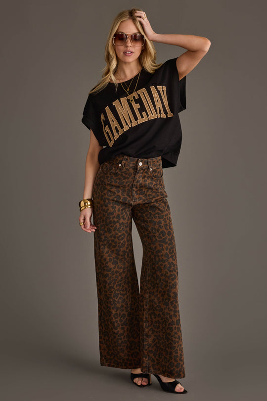 A woman wearing sunglasses, a black & light brown Game Day short sleeve sweatshirt by Panache Apparel Collection, brown leopard-print wide-leg pants, black heels, and gold accessories poses confidently against a plain gray background.