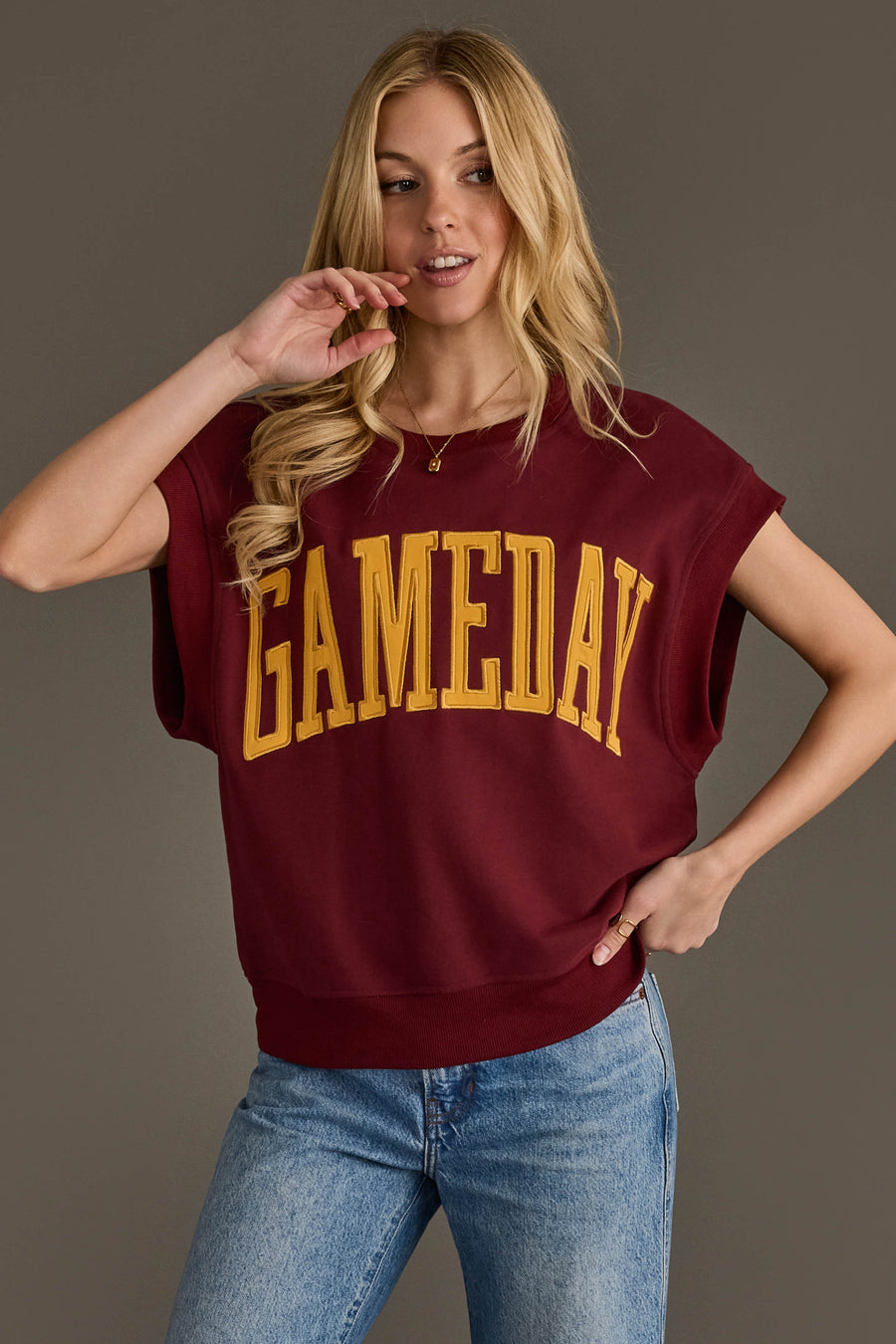 A woman with long blonde hair is wearing the Panache Apparel Collection’s Maroon & Gold Game Day Short Sleeve Sweatshirt and blue jeans, standing against a plain gray background.