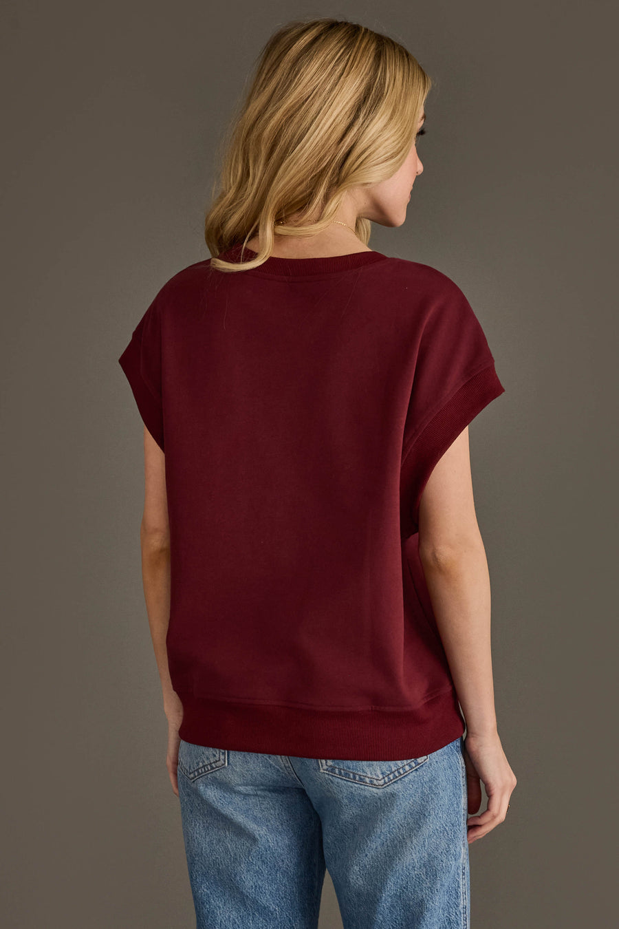 A woman with long blonde hair, wearing the Panache Apparel Collection Maroon & Gold Game Day Short Sleeve Sweatshirt and blue jeans, stands facing away against a plain gray background.