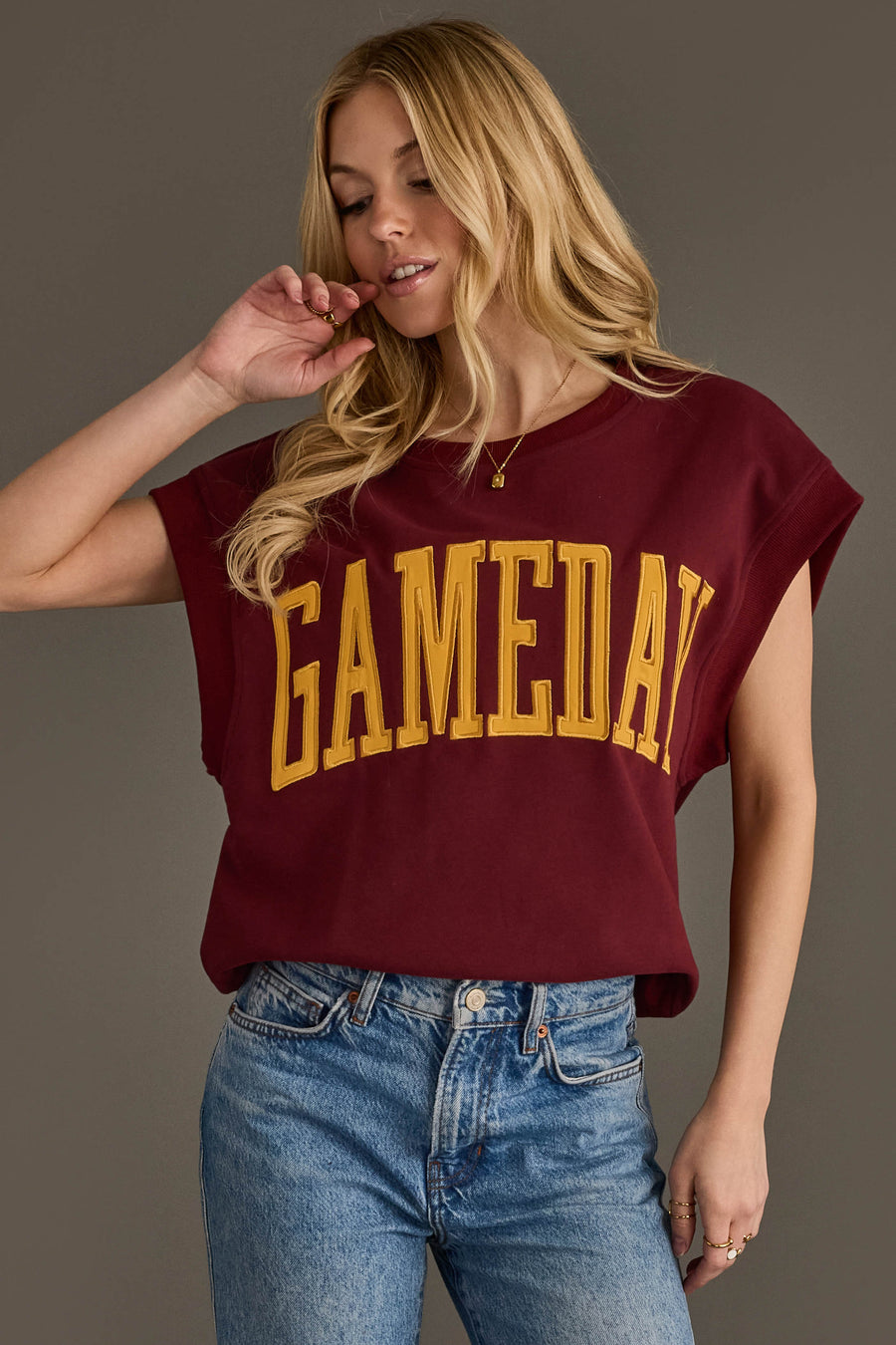 A woman with long blonde hair wears the Maroon & Gold Game Day Short Sleeve Sweatshirt by Panache Apparel Collection, featuring large yellow "GAMEDAY" lettering, paired with blue jeans and standing against a plain background.