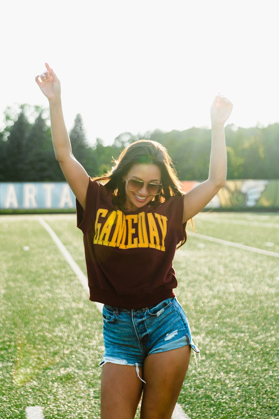 A woman stands on a football field in the sunlight, smiling with arms raised excitedly as she celebrates, wearing sunglasses, denim shorts, and the Maroon & Gold Game Day Short Sleeve Sweatshirt by Panache Apparel Collection.