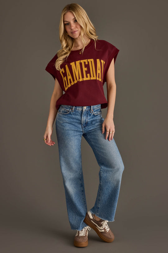 A woman stands against a gray background wearing the Panache Apparel Collection’s Maroon & Gold Game Day Short Sleeve Sweatshirt, paired with blue jeans and brown sneakers. She has long blonde hair and strikes a relaxed pose, with one hand in her pocket.