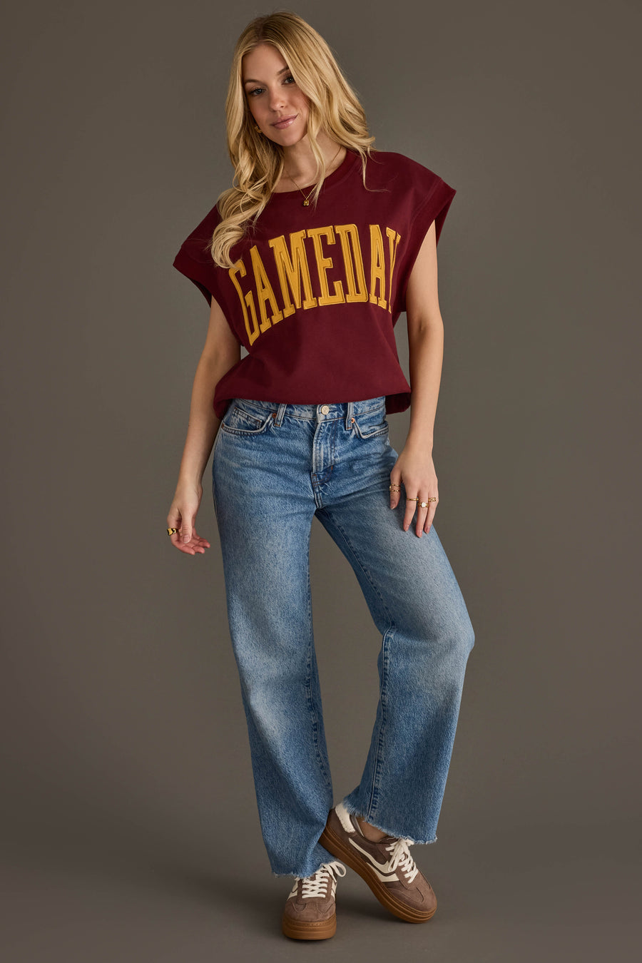 A woman stands against a gray background wearing the Panache Apparel Collection’s Maroon & Gold Game Day Short Sleeve Sweatshirt, paired with blue jeans and brown sneakers. She has long blonde hair and strikes a relaxed pose, with one hand in her pocket.
