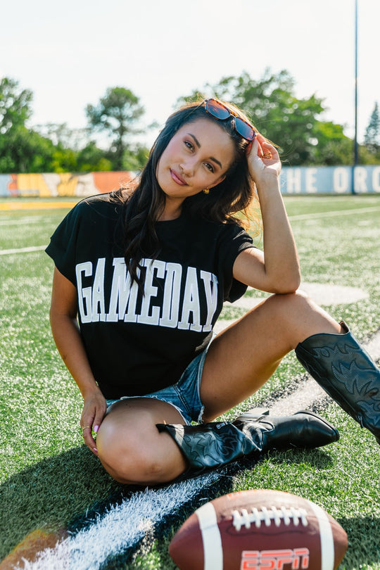 A woman sits on a football field wearing the Panache Apparel Collection Black & White Game Day Short Sleeve Sweatshirt, paired with denim shorts and cowboy boots. She holds sunglasses on her head, with a football in the foreground.