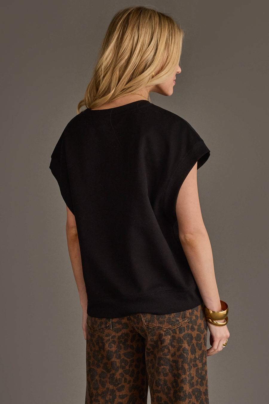 A woman with long blonde hair faces away, wearing the Panache Apparel Collection's Black & White Game Day Short Sleeve Sweatshirt and brown leopard print pants. She accessorizes with gold bangles on her right wrist, and the background is plain and neutral.