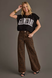 A smiling woman in a Black & White Game Day Short Sleeve Sweatshirt by Panache Apparel Collection, wide-leg brown leopard print pants, black heels, and gold jewelry poses against a plain gray background.