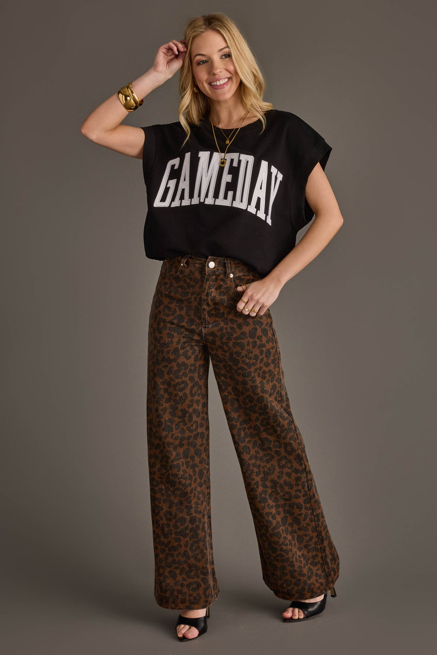 A smiling woman in a Black & White Game Day Short Sleeve Sweatshirt by Panache Apparel Collection, wide-leg brown leopard print pants, black heels, and gold jewelry poses against a plain gray background.