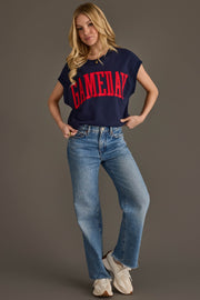 A woman with long blonde hair poses with one hand on her hip against a plain background, wearing the Panache Apparel Collection’s Navy & Red Game Day Short Sleeve Sweatshirt, light blue jeans, and white sneakers. GAMEDAY is featured in bold red letters across the front of the sweatshirt.