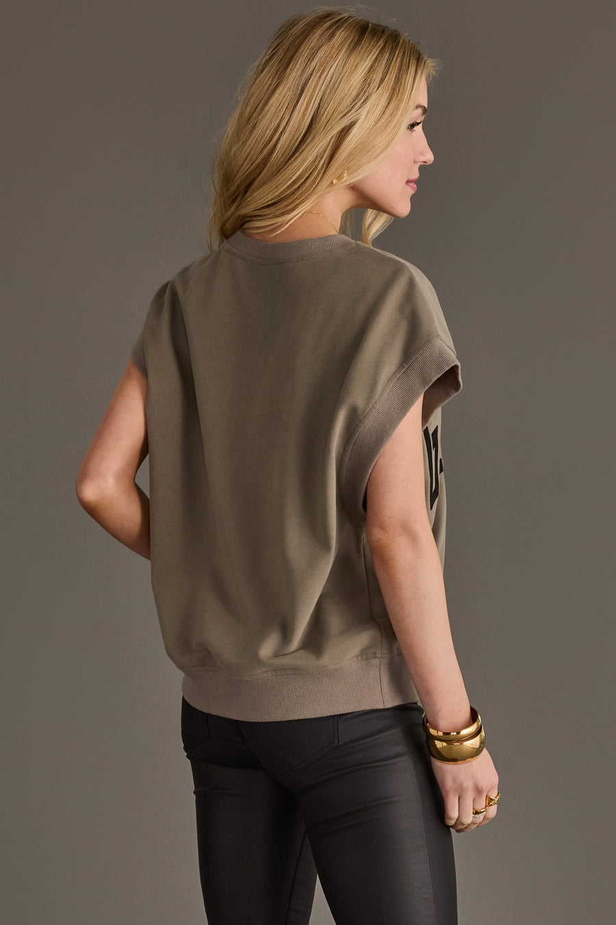 A woman with long blonde hair stands sideways, facing away. She wears the Panache Apparel Collection Gray/Olive & Black Game Day Short Sleeve Sweatshirt and black pants, accessorized with gold bangles. The background is plain and neutral.