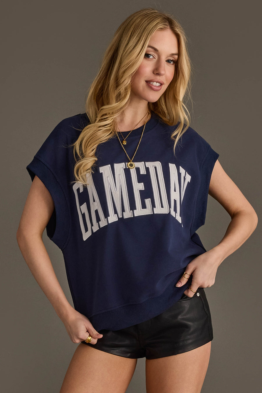 A woman with long blonde hair smiles and holds the hem of her Panache Apparel Collection Navy & White Game Day Short Sleeve Sweatshirt, which has "GAMEDAY" in large white letters, paired with black leather shorts.