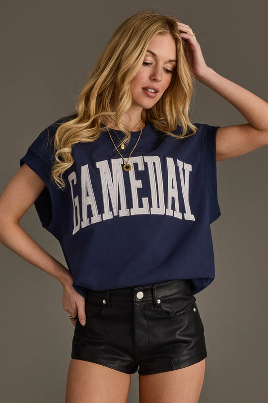 A young woman with long blonde hair wears the Panache Apparel Collection Navy & White Game Day Short Sleeve Sweatshirt paired with black leather shorts, standing with one hand on her hip and the other touching her hair.