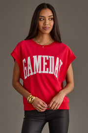 A woman stands with her hands together against a plain gray background, wearing the Panache Apparel Collection Red & White Game Day Short Sleeve Sweatshirt with bold white "GAMEDAY" lettering, accessorized with gold jewelry.