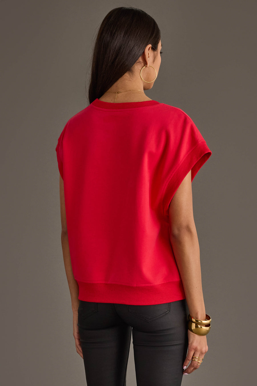 A woman with long dark hair stands facing away, wearing the Panache Apparel Collection Red & White Game Day Short Sleeve Sweatshirt, paired with black pants, large hoop earrings, and gold bracelets against a plain gray background.