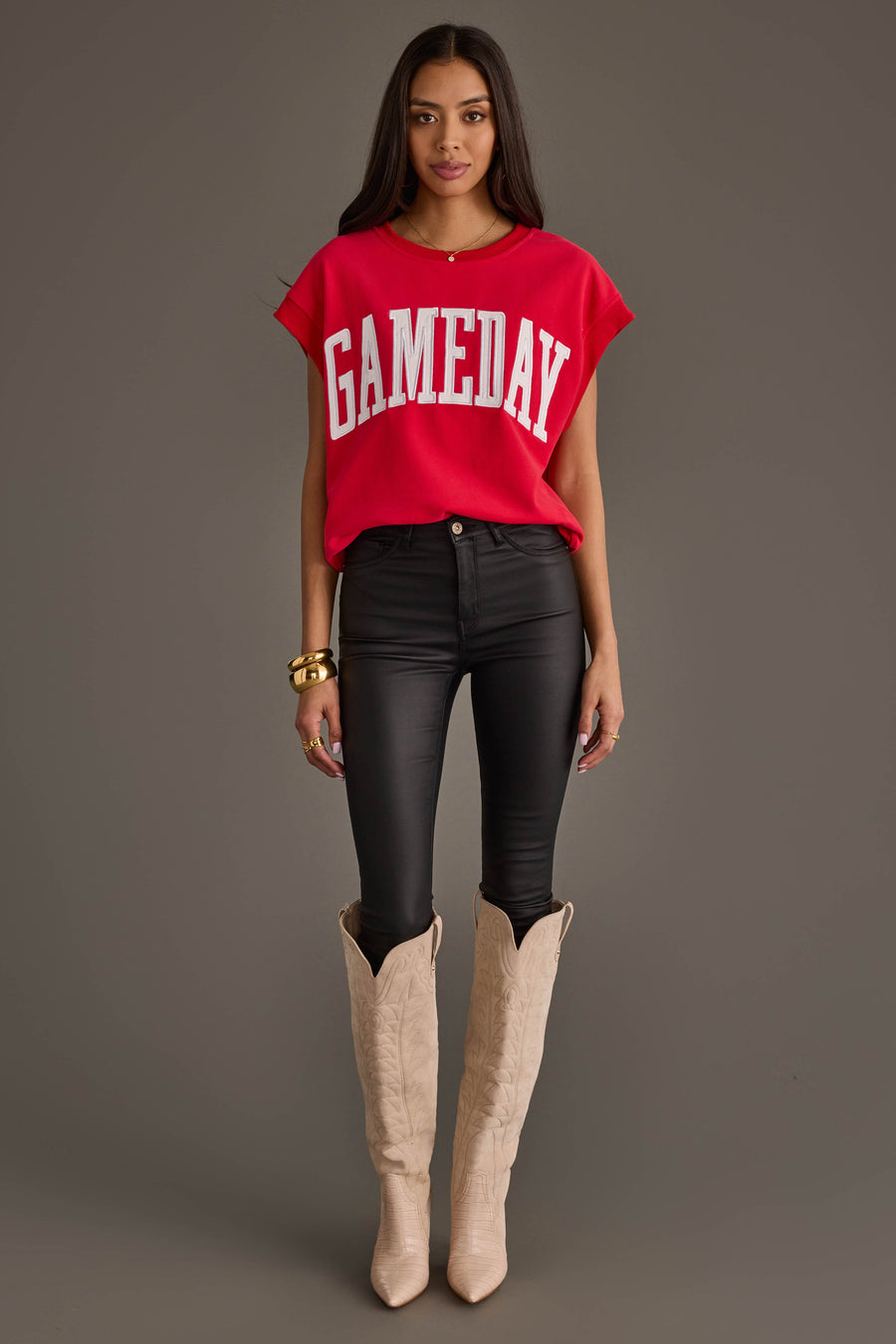 A woman stands against a plain gray background wearing the Panache Apparel Collection Red & White Game Day Short Sleeve Sweatshirt, black skinny jeans, tan cowboy boots, and gold bracelets.