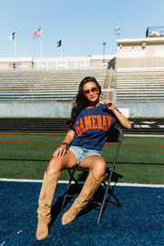 A woman in sunglasses and a relaxed fit Navy & Orange Game Day Short Sleeve Sweatshirt from the Panache Apparel Collection sits on a chair on a football field, wearing denim shorts and tan boots. Bleachers, flags, and a clear sky are visible in the background.
