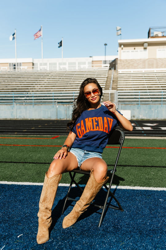 A woman in sunglasses and a relaxed fit Navy & Orange Game Day Short Sleeve Sweatshirt from the Panache Apparel Collection sits on a chair on a football field, wearing denim shorts and tan boots. Bleachers, flags, and a clear sky are visible in the background.