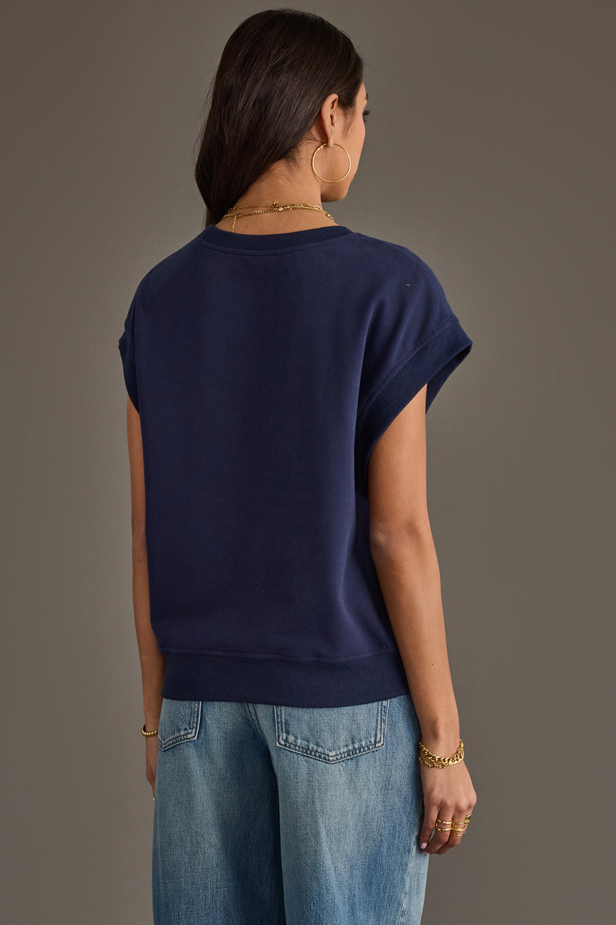 A woman with long dark hair is seen from behind wearing the Panache Apparel Collection Navy & Orange Game Day Short Sleeve Sweatshirt paired with light blue jeans. She completes her look with gold hoop earrings, bracelets, and a necklace.