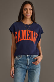 A woman wearing the Panache Apparel Collection Navy & Orange Game Day Short Sleeve Sweatshirt, featuring bold orange "GAMEDAY" lettering, paired with blue jeans and gold jewelry, smiles against a plain background.