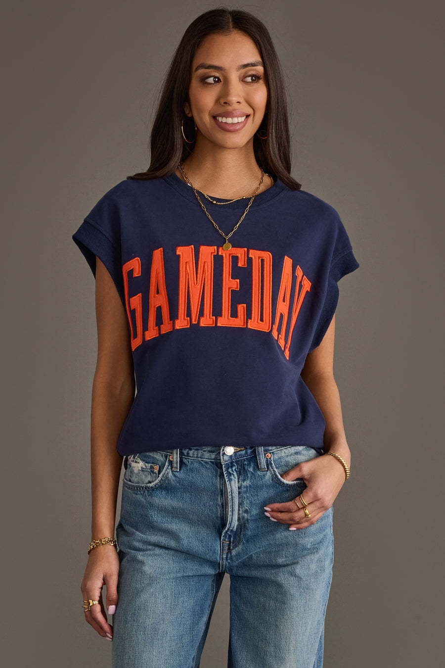 A woman wearing the Panache Apparel Collection Navy & Orange Game Day Short Sleeve Sweatshirt, featuring bold orange "GAMEDAY" lettering, paired with blue jeans and gold jewelry, smiles against a plain background.