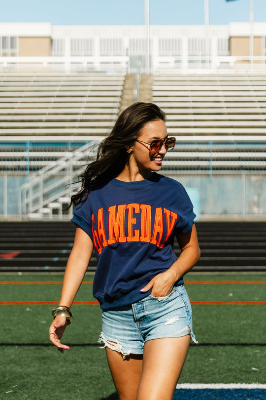 A woman wearing sunglasses, denim shorts, and the Panache Apparel Collection Navy & Orange Game Day Short Sleeve Sweatshirt stands on a sports field with empty bleachers in the background.