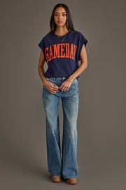 A woman stands against a plain background wearing a Panache Apparel Collection Navy & Orange Game Day Short Sleeve Sweatshirt, light blue flared jeans, and platform shoes. She has long dark hair and minimal accessories.