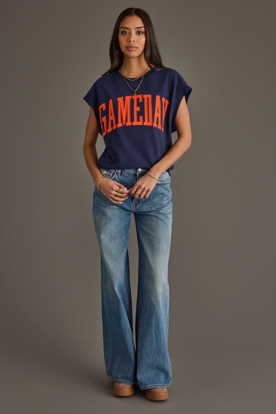 A woman stands against a plain background wearing a Panache Apparel Collection Navy & Orange Game Day Short Sleeve Sweatshirt, light blue flared jeans, and platform shoes. She has long dark hair and minimal accessories.