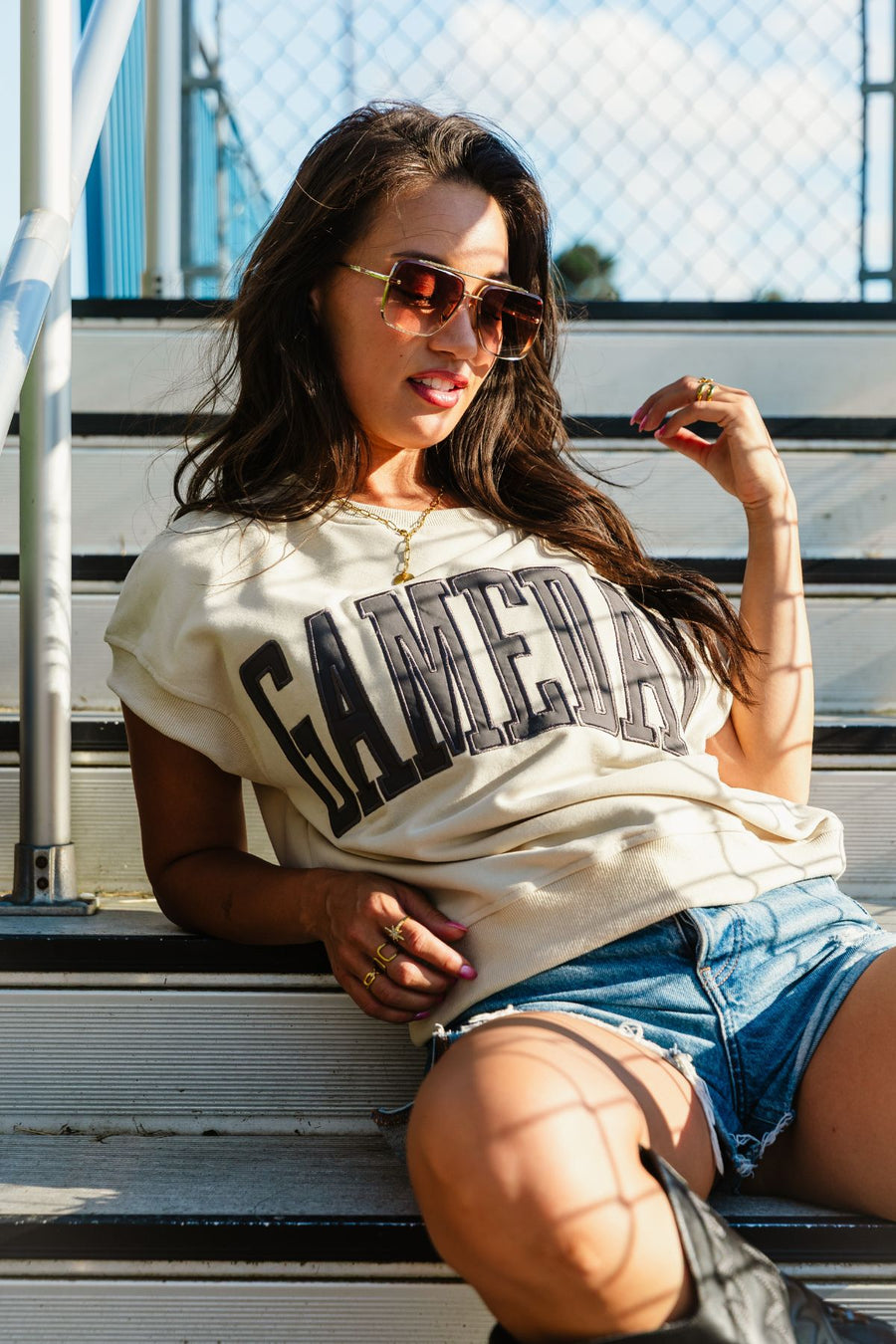 A woman with long hair sits relaxed on outdoor bleachers, sunlight and shadows playing across her as she poses with one hand near her face. She wears sunglasses, denim shorts, boots, and the Beige & Gray Game Day Short Sleeve Sweatshirt from Panache Apparel Collection.
