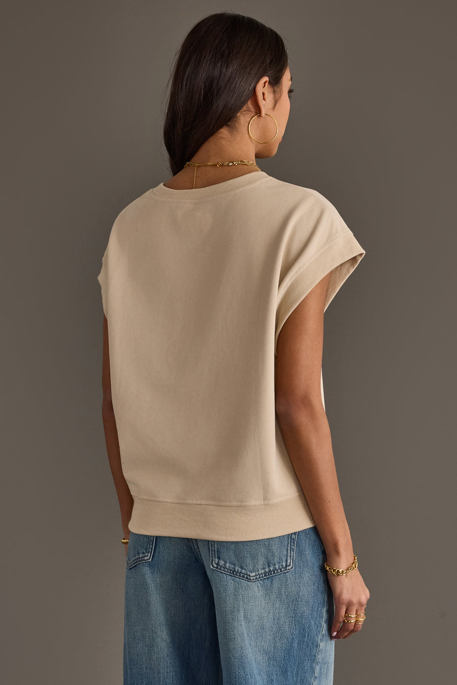 A woman with long dark hair wears the Panache Apparel Collection Beige & Gray Game Day Short Sleeve Sweatshirt, along with blue jeans, gold hoop earrings, and bracelets. She stands facing away from the camera against a plain brown background.