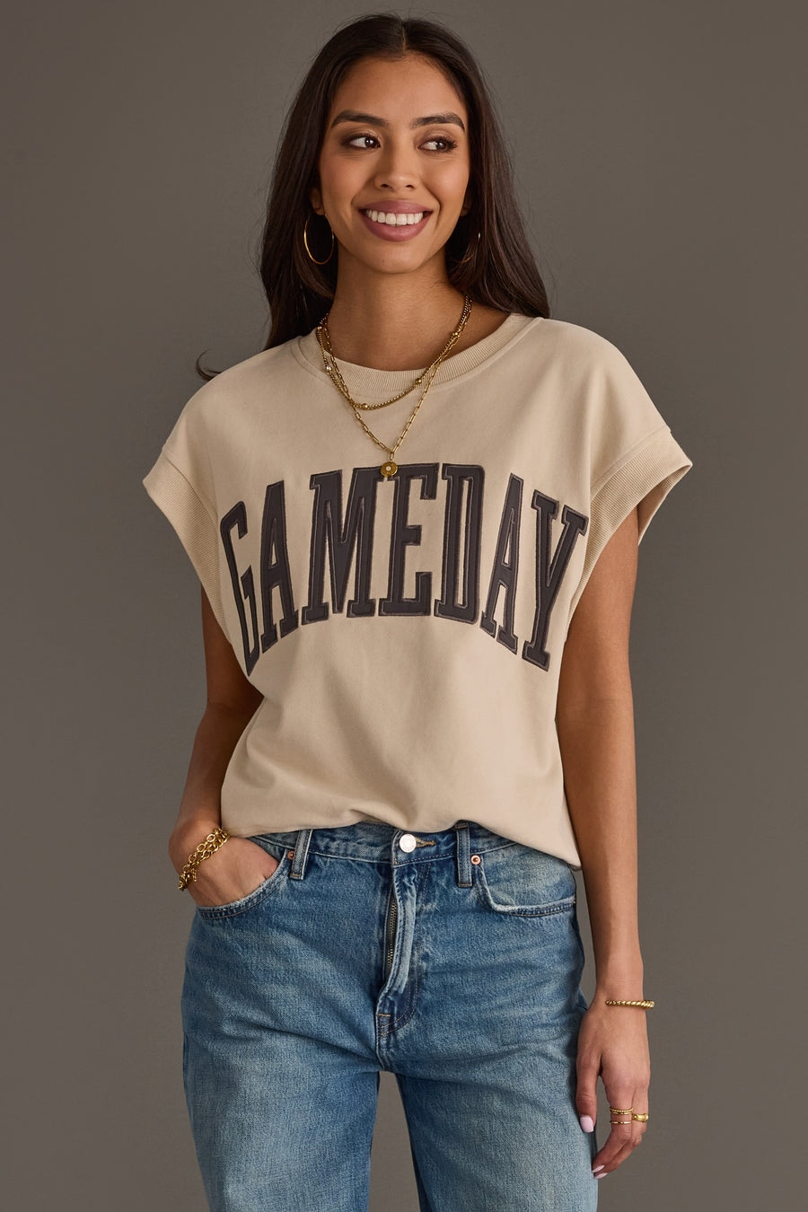 A woman wearing a Beige & Gray Game Day Short Sleeve Sweatshirt from Panache Apparel Collection, blue jeans, gold necklaces, hoop earrings, and bracelets stands smiling with one hand in her pocket against a gray background.